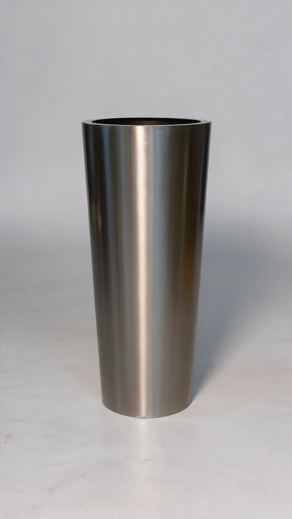 A brushed stainless steel cylinder, likely a tumbler, vase, or planter, standing upright on a light gray surface against a neutral gray background.